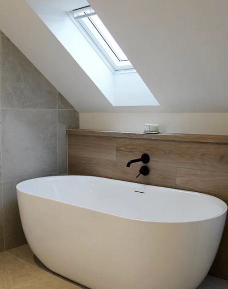 A simple white bath sits in a grey tiled bathroom under the eaves